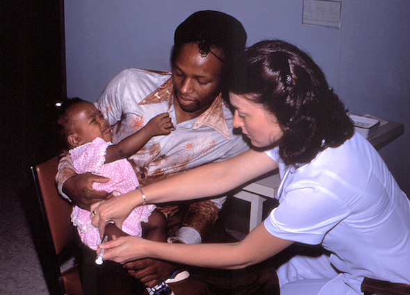 A father holds his daughter as she receives childhood vaccinations (Dekalb County, Ga., 1977).